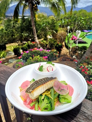A white plate with a grilled fish, vegetables, and a small bowl of sauce, set against a garden backdrop with palm trees and flowers.