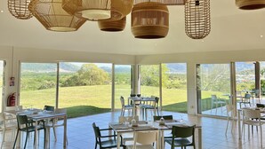 A spacious dining area with a table set for four, large windows offering a view of a green landscape, and multiple hanging wicker chandeliers. - Rivland Resort (Paita)