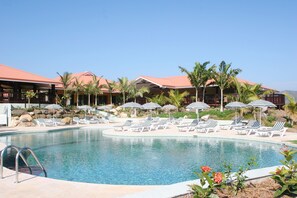 A resort pool area with lounge chairs and umbrellas, surrounded by palm trees and a building with a red roof.