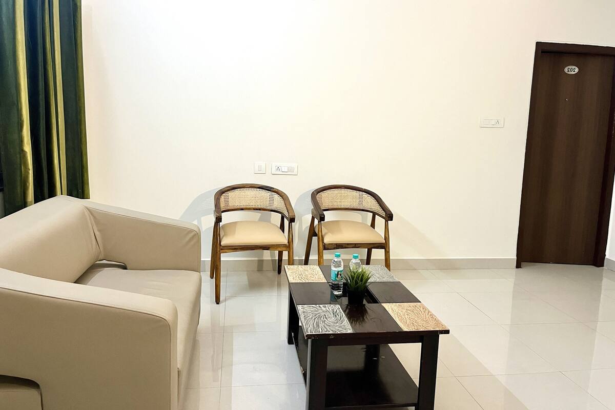 A room with a beige sofa, two wooden chairs, a small table with a plant and water bottles, and a door with a number plate.