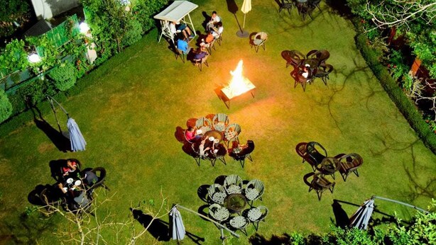 Ein Sitzbereich im Freien mit Tischen und Stühlen, die um eine Feuerstelle angeordnet sind, umgeben von Pflanzen und einer Hecke.