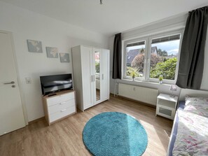 A room with a bed, a TV on a wooden cabinet, a large window with curtains, and a blue rug.