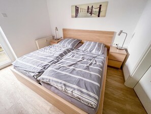 A modern bedroom with a wooden bed frame, a nightstand, a lamp, and a painting on the wall. - Apartment house Nige Ooge Apartment No. 1 - Apartment with small terrace (Cuxhaven)