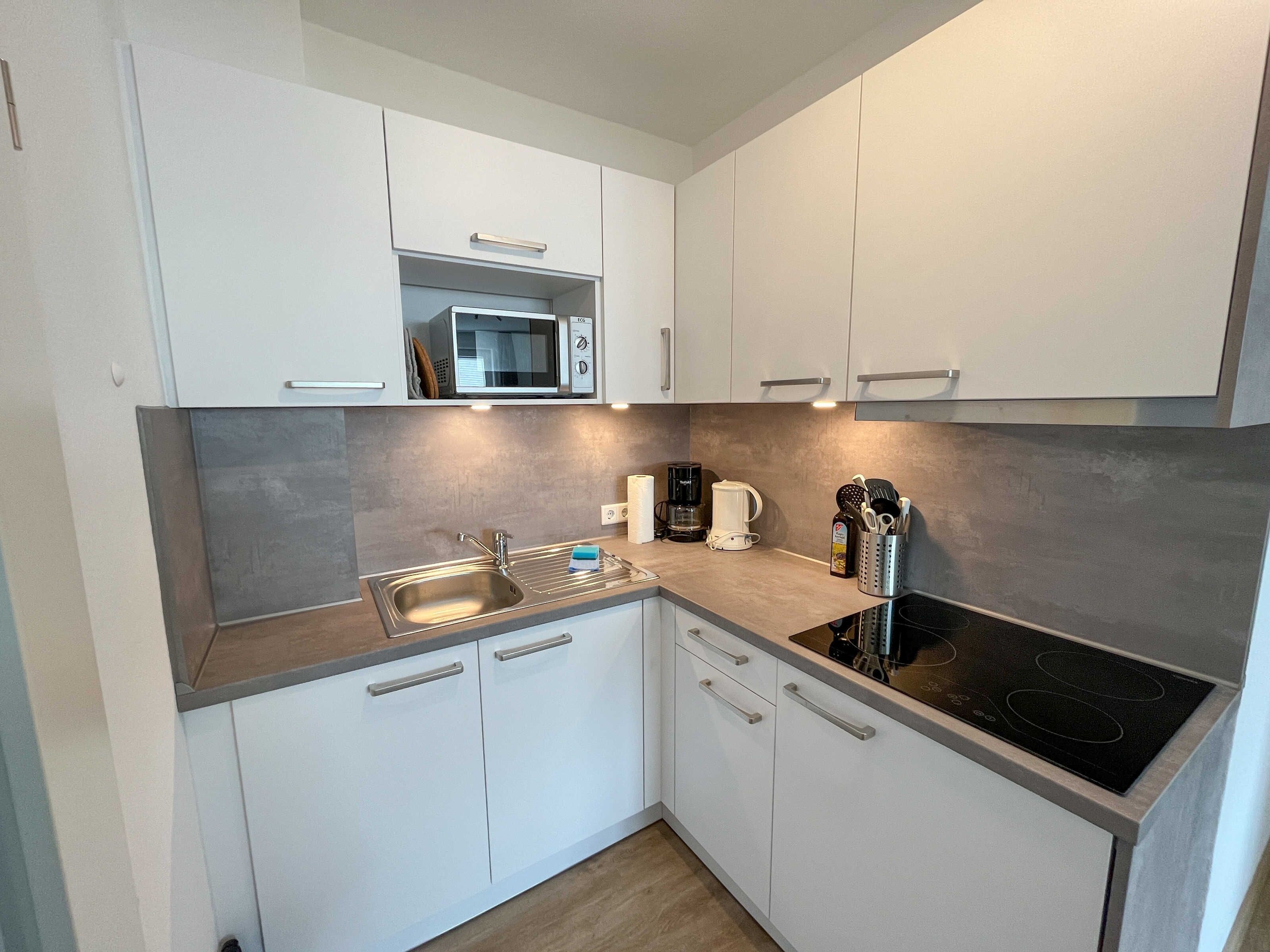 Una cocina moderna con armarios blancos, un lavamanos de acero inoxidable, un microondas, una cafetera y una placa de inducción negra.