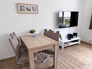 A modern room with a wooden dining table, two chairs, a small potted plant, a white shelf with a TV, speakers, and a remote control. - Apartment house Nige Ooge Apartment No. 1 - Apartment with small terrace (Cuxhaven)