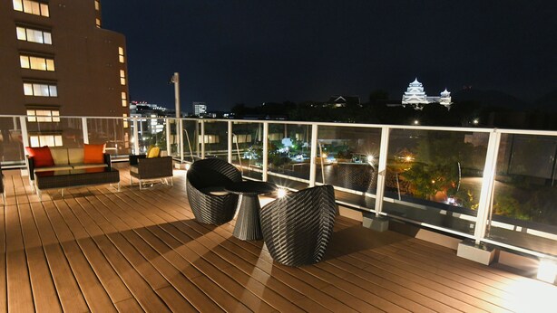 A wooden deck with outdoor seating, a glass railing, and a view of a castle-like building at night. - &and COMFY HOTEL Kumamotojo View (Kumamoto)