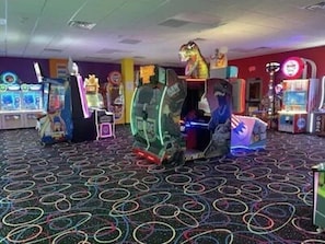 Huoneessa on pelihalleihin tyypillisiä peliautomaatteja, pyöreäkuvioinen matto ja hyllyllä seisova dinosauruksen patsas.