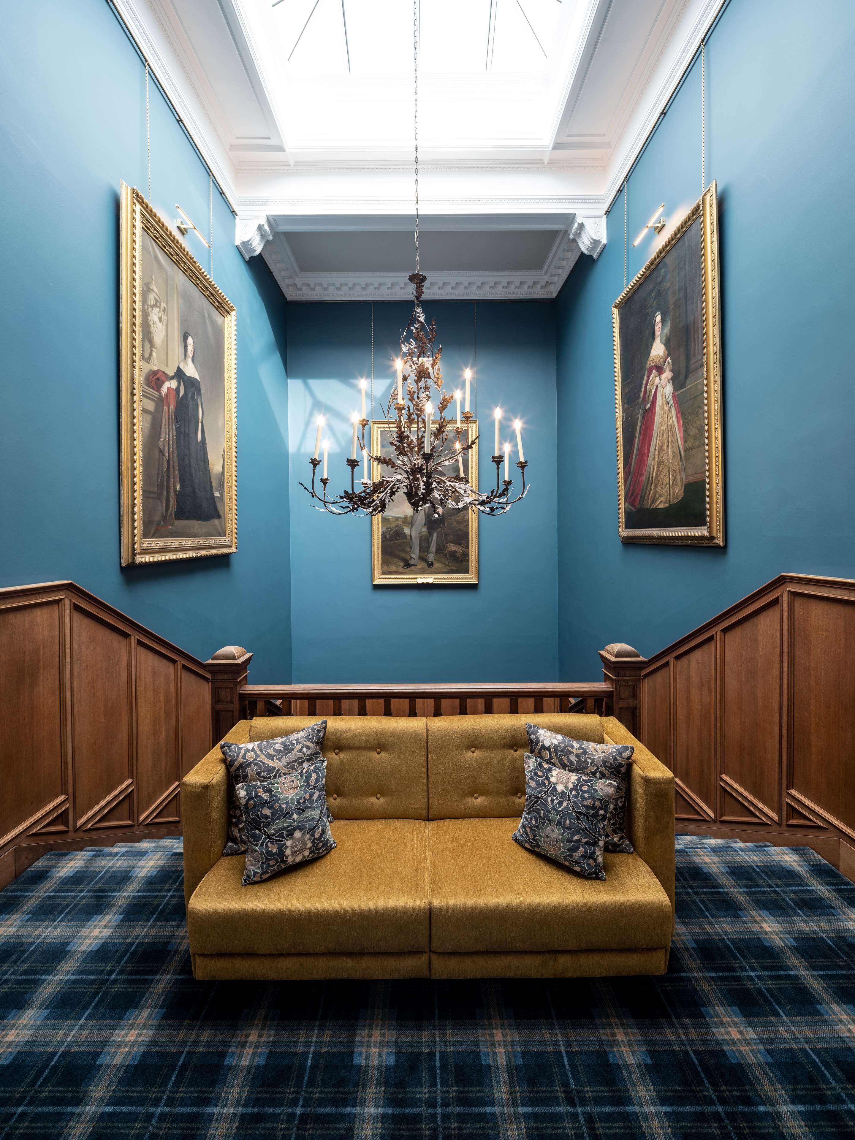 A room with a blue wall, a chandelier, a sofa with patterned cushions, and two framed paintings.