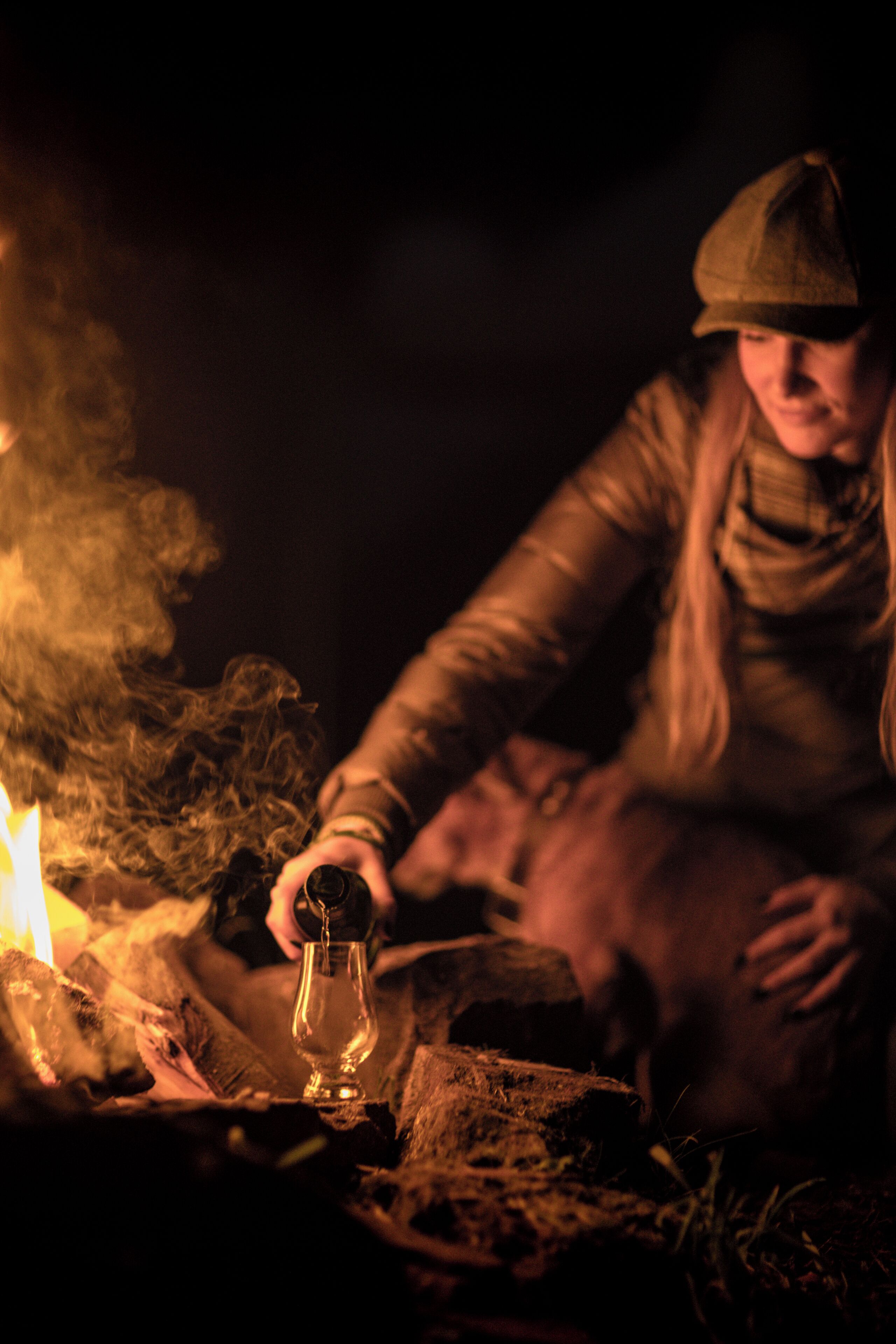 A person sitting by a campfire, holding a glass.