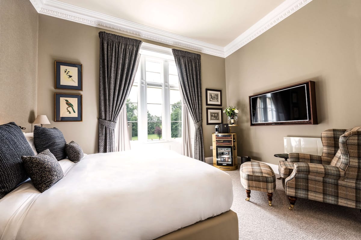 A hotel room with a large bed, a television mounted on the wall, a seating area with a chair and ottoman, and framed pictures on the wall.