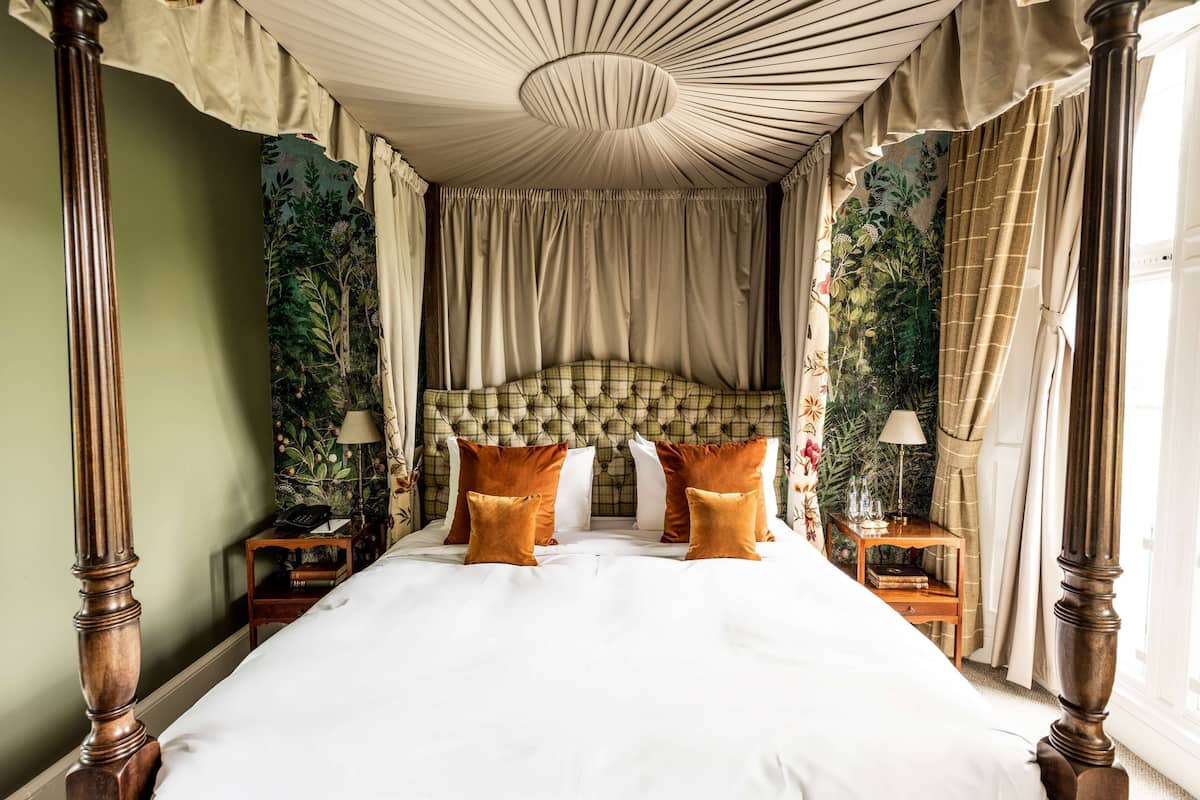 A four-poster bed with a tufted headboard, two nightstands with lamps, and a green wall with a floral tapestry.