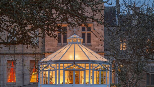 A conservatory with a glass roof, surrounded by a gravel path and lit lanterns, leading to a building with a stone facade.