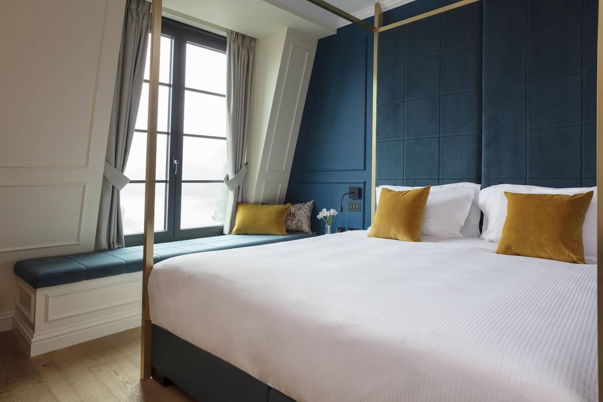 A bedroom with a large bed, a window with curtains, a bench, and a blue accent wall.