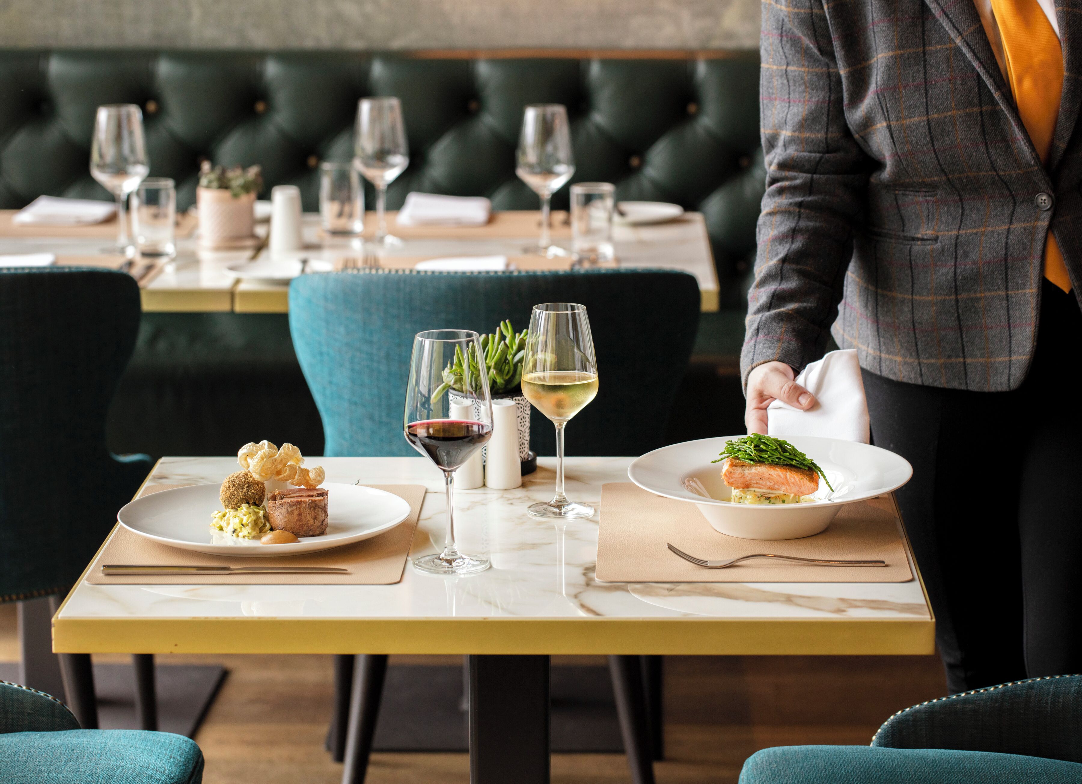 Ein Restaurantambiente mit einem Tisch für zwei Personen, bestecktem Tisch, einem Teller mit Speisen, einem Glas Rotwein und einem Glas Weißwein.