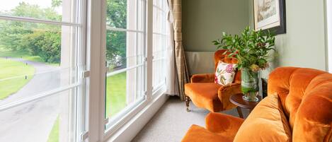 A room with a large window, a view of a road and greenery, a plush orange sofa, and a small table with a vase of flowers.