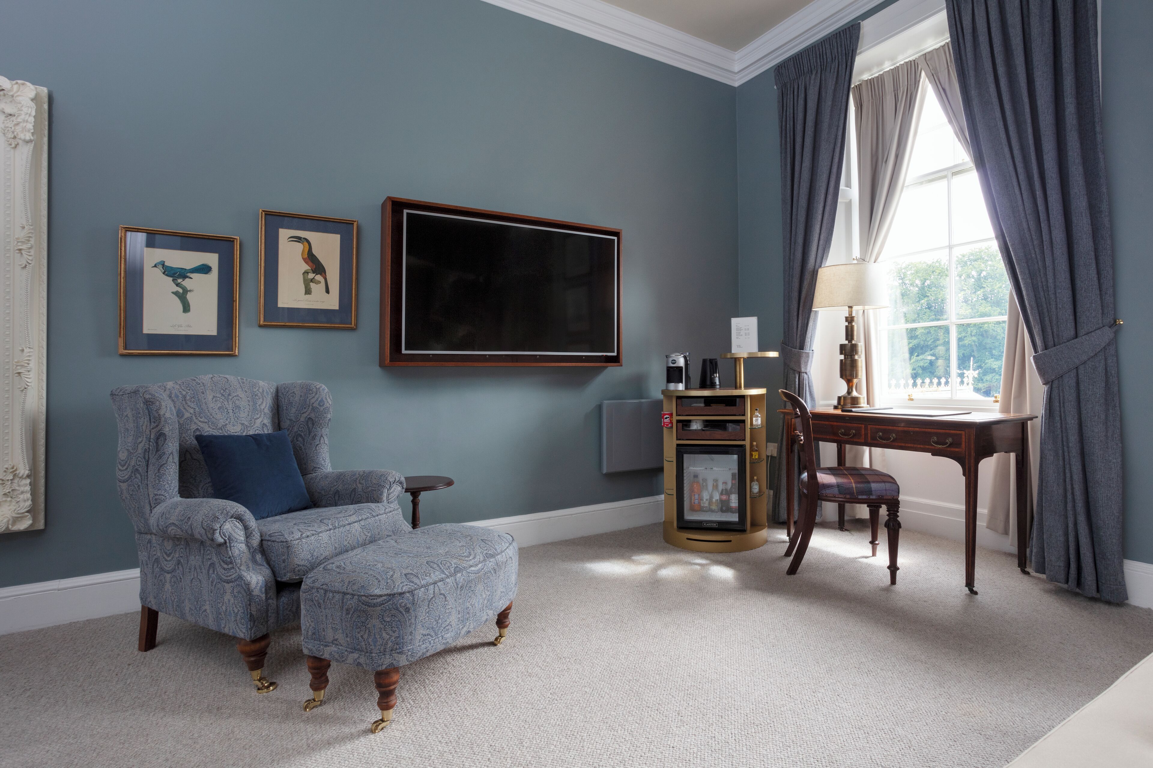A room with a patterned armchair, a desk with a lamp, and framed artwork on the wall.