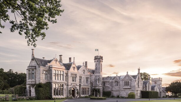A large, historic stone building with multiple towers and a central entrance, surrounded by well-maintained lawns and a paved driveway.