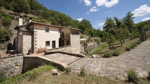 Una casa de piedra con un puente de piedra que lleva a un patio, rodeado de árboles y colinas.