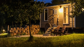 Een klein houten huisje met een kruiwagen, twee houten stoelen en een stapel hout.