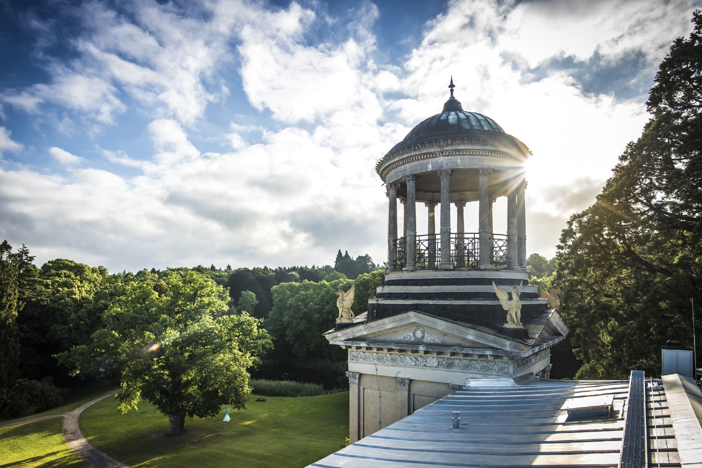 A classical pavilion with a dome and columns, surrounded by lush greenery and a clear sky.