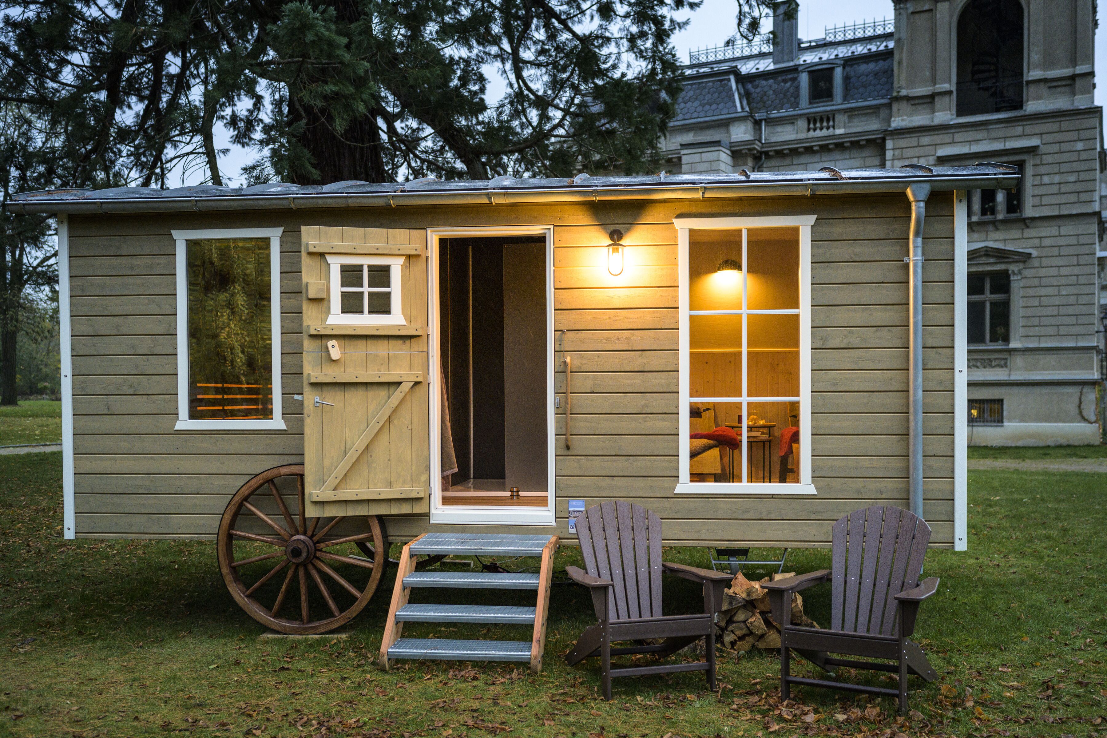 A small wooden cabin with a porch, a large wheelbarrow, and two Adirondack chairs.