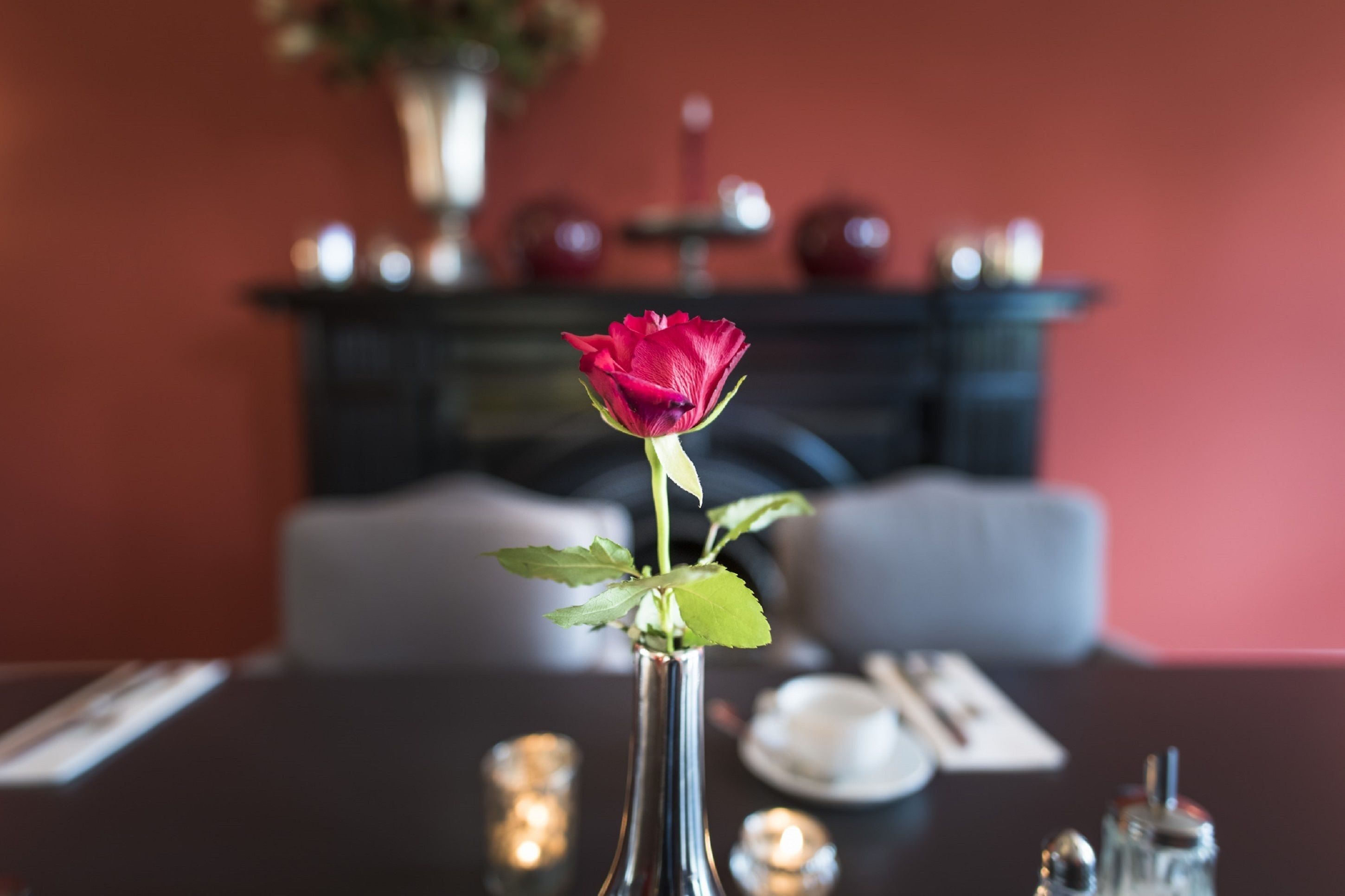 A dining table with a vase containing a single red rose, candles, and a plate with a knife and fork.