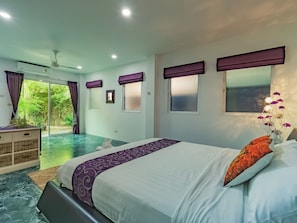 A bedroom with a bed, a dresser, a window with curtains, and a view of a garden.