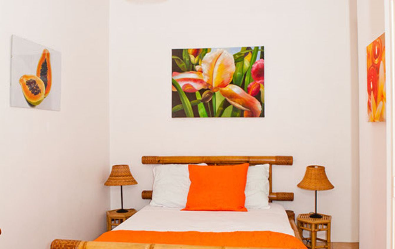 Quarto de hotel com cama, duas luminárias de cabeceira, um quadro floral pendurado na parede e uma pequena imagem de frutas.