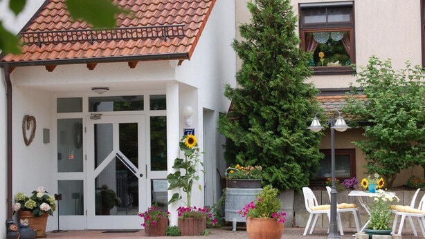A building with a tiled roof, a glass door entrance, and a small patio area with outdoor seating and potted plants. - Landhotel Mühlberg (Moessingen)