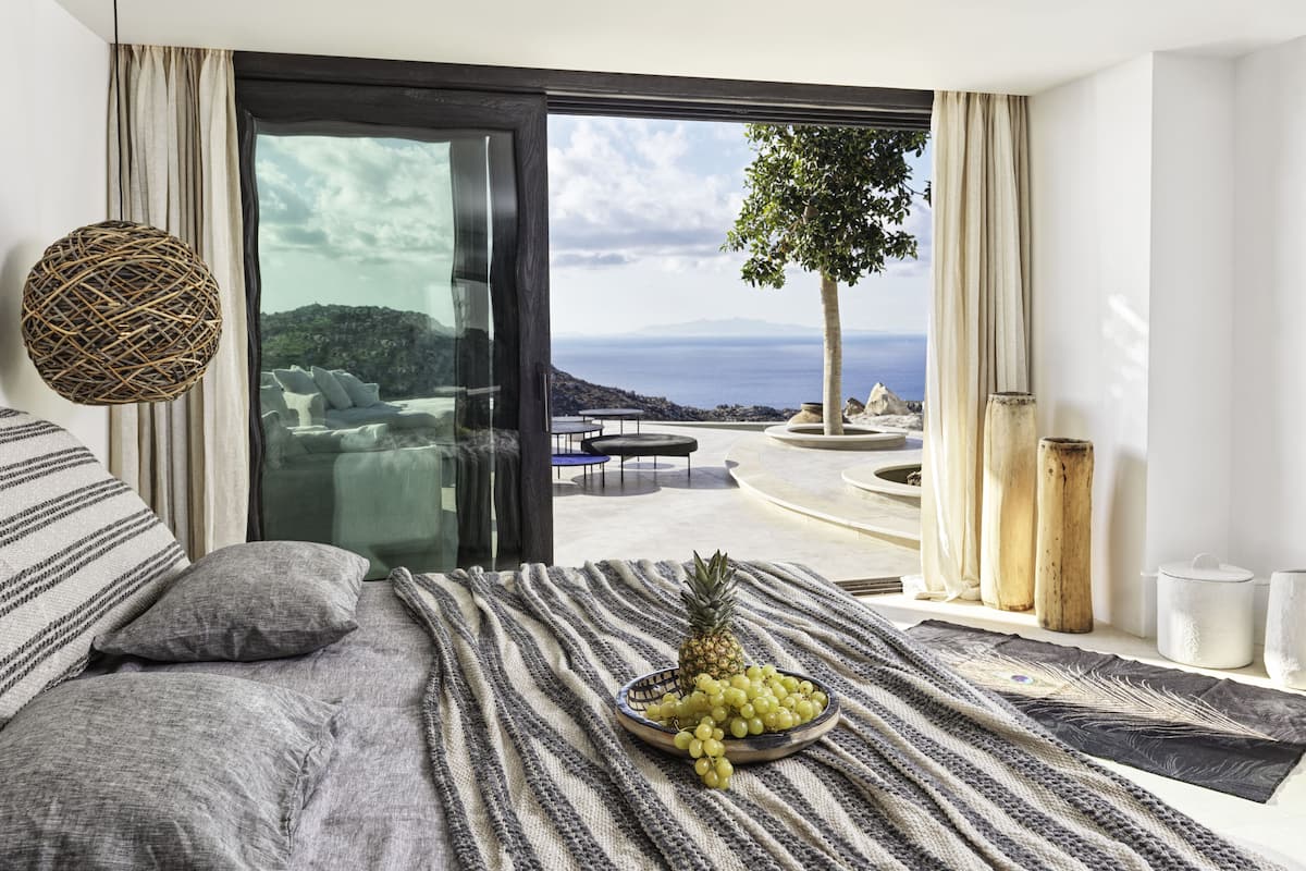 A bedroom with a large window, a bed with a striped blanket, a bowl of fruit, and a view of the sea and mountains.
