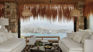 A room with a white sofa, a round coffee table with a bowl of fruit, and a view of a coastal landscape. - Myconian Panoptis Escape - Small Luxury Hotels of the World (Mykonos)