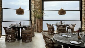 A dining area with wicker chairs and tables, large windows with a view, and hanging wicker light fixtures. - Myconian Panoptis Escape - Small Luxury Hotels of the World (Mykonos)