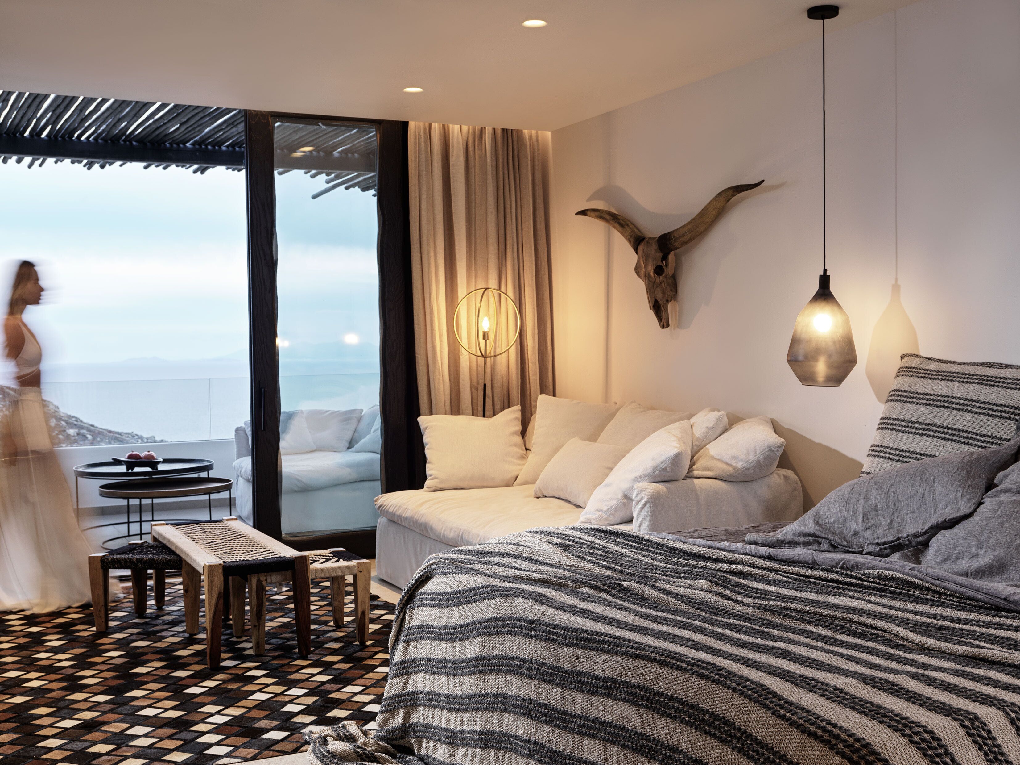 A modern hotel room with a large bed, a sofa, a small table, and a view of the ocean.