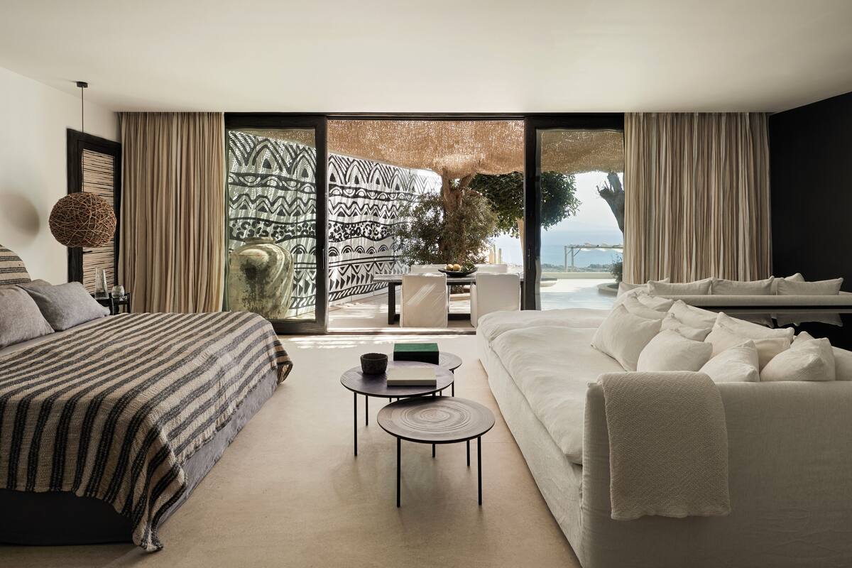 A modern hotel room with a large bed, a sofa, a small round table, and a view of the sea through large glass doors.