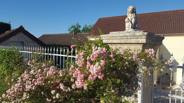 A stone lion statue atop a building with a tiled roof, surrounded by blooming pink roses. - Maison D'Hote Le Relais (Fresnes)