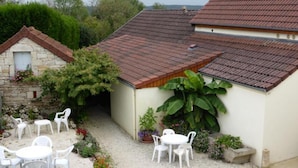 A building with a tiled roof, a stone structure with a door, and a garden area with white plastic chairs and tables. - Maison D'Hote Le Relais (Fresnes)