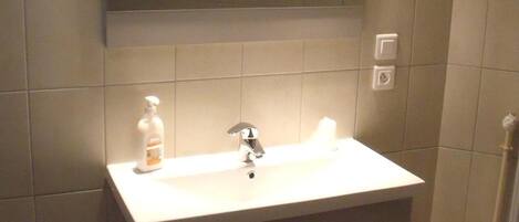 Un baño con lavamanos blanco, espejo y armarios. Hay un cubo de basura y una botella de jabón para manos sobre el lavamanos.