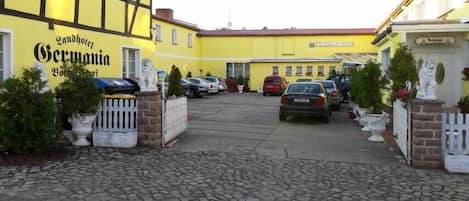 A hotel entrance with a cobblestone driveway, parked cars, and a yellow building with a sign reading "Landhotel Germanyia".
