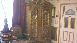 Un cabinet en bois richement décoré, orné de sculptures complexes, un fauteuil ancien et un tapis rouge menant à une porte dotée d’un cadre doré.