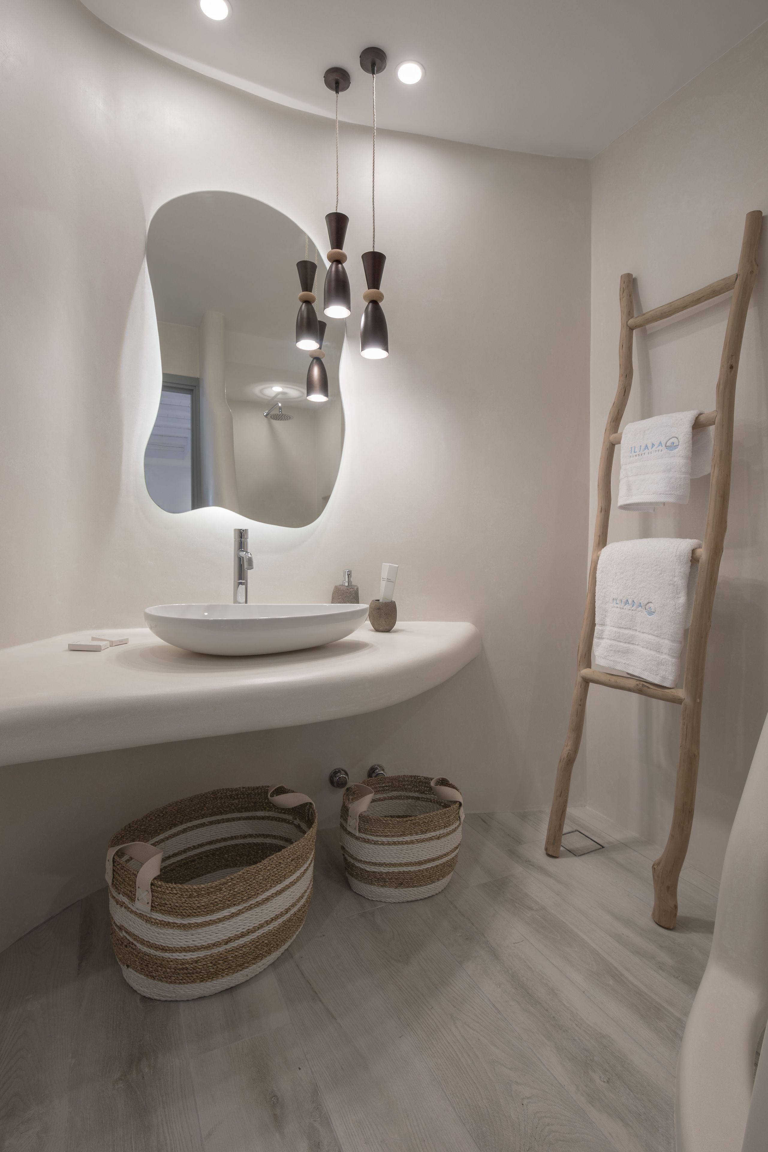 A modern bathroom with a white sink, a large mirror, and hanging pendant lights. There are two woven baskets on the floor and a wooden ladder with a towel hanging on it.