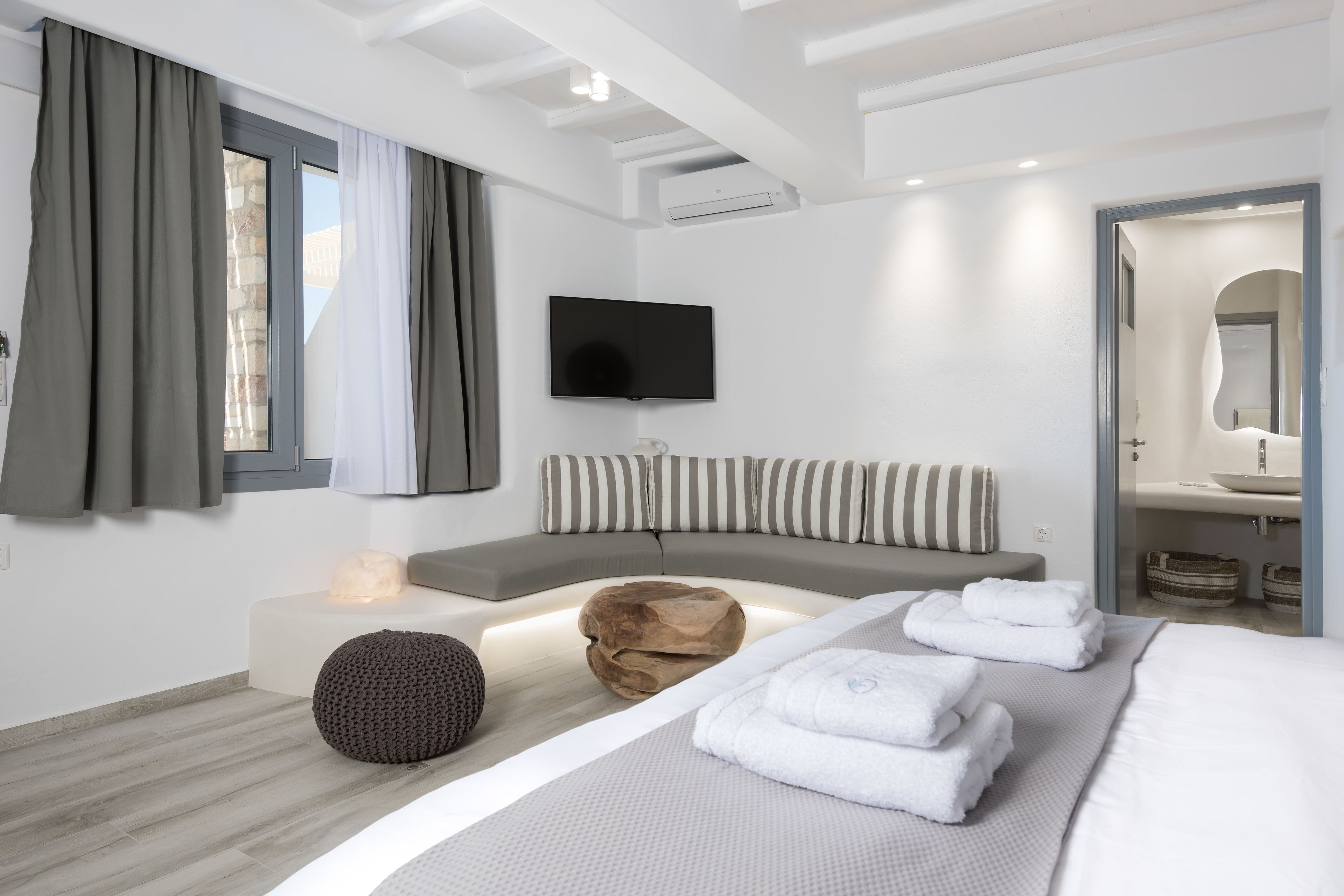 A modern hotel room with a large bed, a seating area, a flat-screen TV, and a bathroom with a mirror and sink.