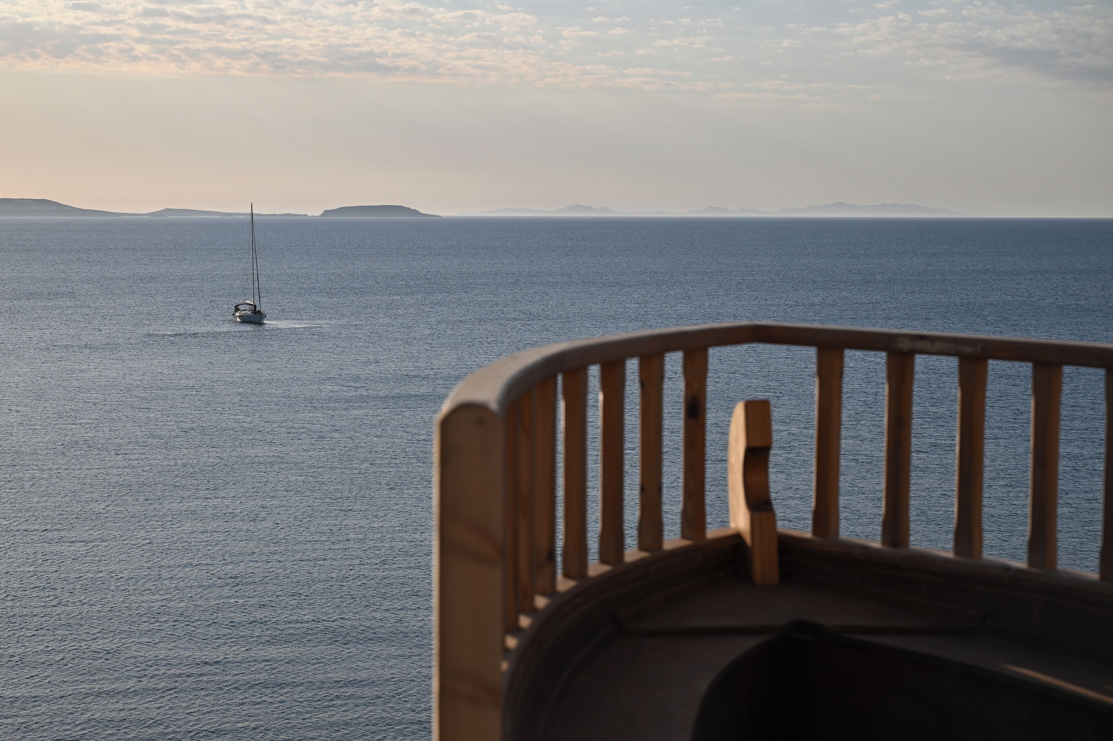 Une vue sur la mer depuis un balcon dot é d’une balustrade en bois, avec un voilier à l’horizon et une terre lointaine.