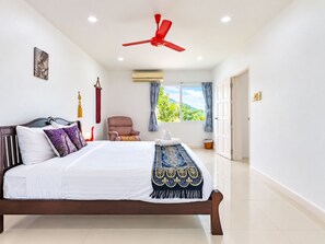 A bedroom with a bed, a ceiling fan, a chair, a window with curtains, and a view of greenery outside. - Private Pool Villa for your family and friends (Tambon Chalong)