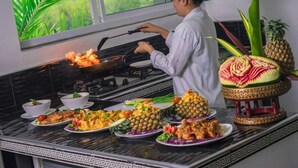 A chef cooking on a stove with a view of greenery outside, surrounded by various dishes and a decorative watermelon centerpiece. - Private Pool Villa for your family and friends / Private Chef available (Tambon Chalong)