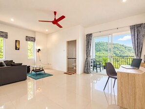 A modern living room with a sofa, a dining table with chairs, a red ceiling fan, and a balcony with a view of a forested hill. - Private Pool Villa for your family and friends / Private Chef available (Tambon Chalong)