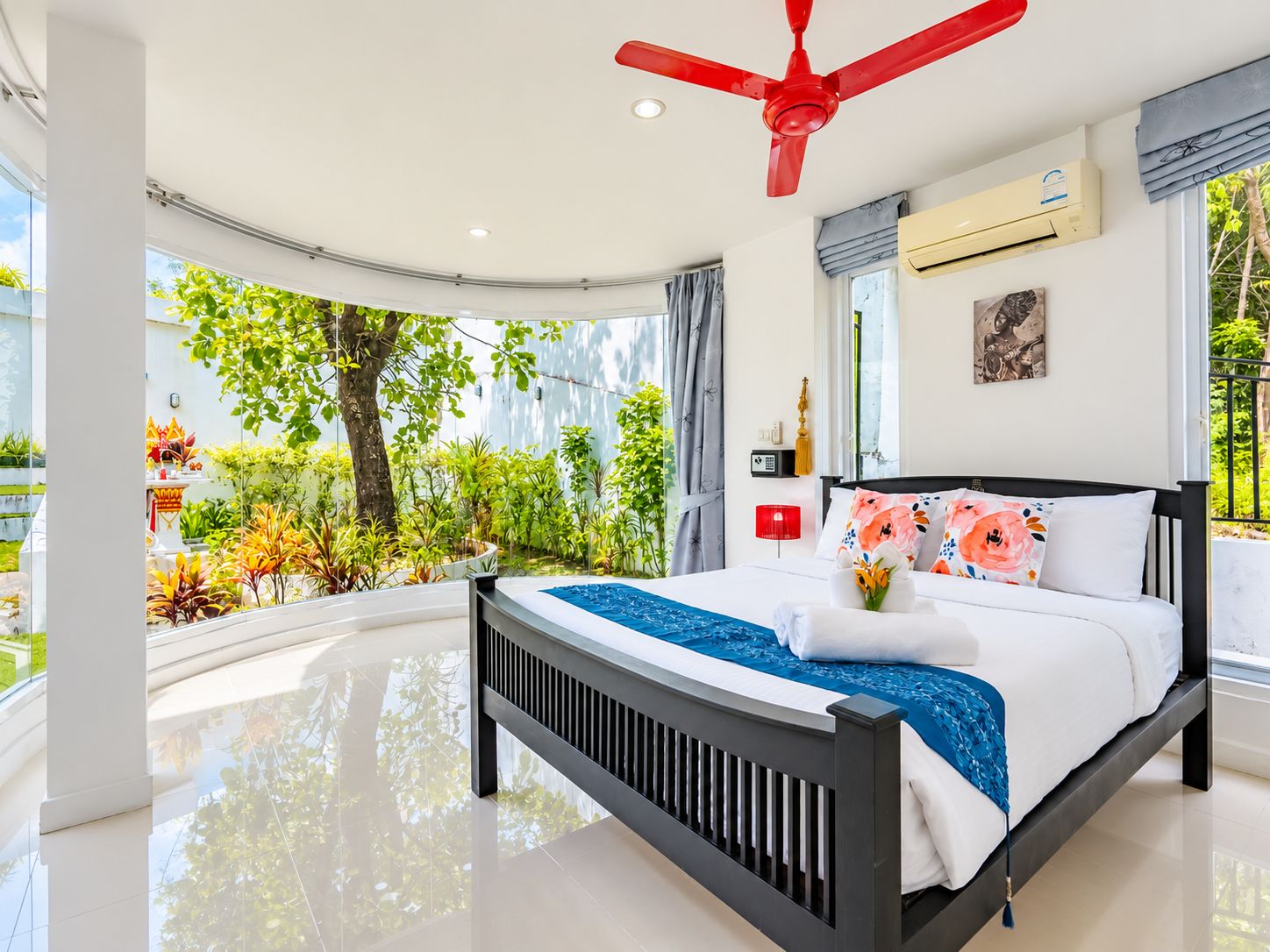 Ein modernes Schlafzimmer mit gewölbter Decke, einem Bett mit schwarzem Gestell, weißer Bettwäsche und einem blauen Akzent. Es gibt einen roten Deckenventilator, eine Klimaanlage und einen Blick auf Grünfläche vor dem Fenster.