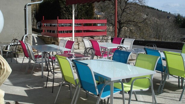An outdoor seating area with tables and chairs, a red awning, and a stone wall in the background.