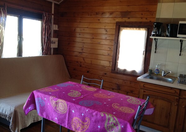 A cozy cabin interior with a wooden interior, a dining table with a patterned tablecloth, a sofa, a window with curtains, and a kitchen area with a sink and appliances.