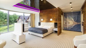 Un dormitorio moderno con una cama grande, un sillón blanco y vistas a una casa a través de amplios ventanales.