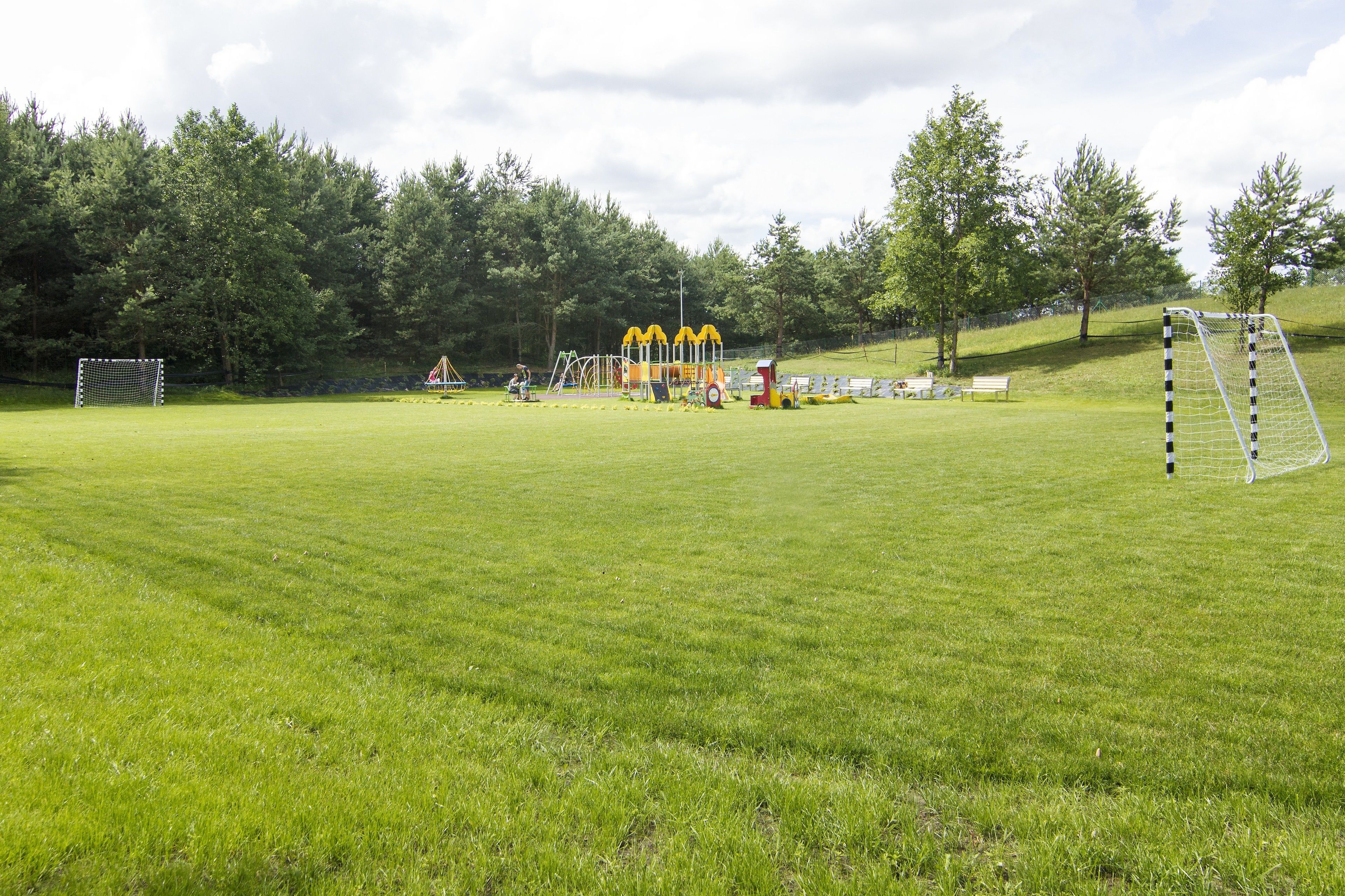 Un terrain herbeux bien entretenu, équipé de deux buts de football, une aire de jeux avec des structures d’escalade jaunes et un espace avec des bancs.